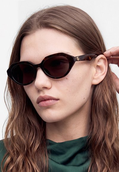 Jeune femme aux longs cheveux bruns portant des lunettes de soleil sombres et angulaires et un top vert foncé, ajustant la branche des lunettes.