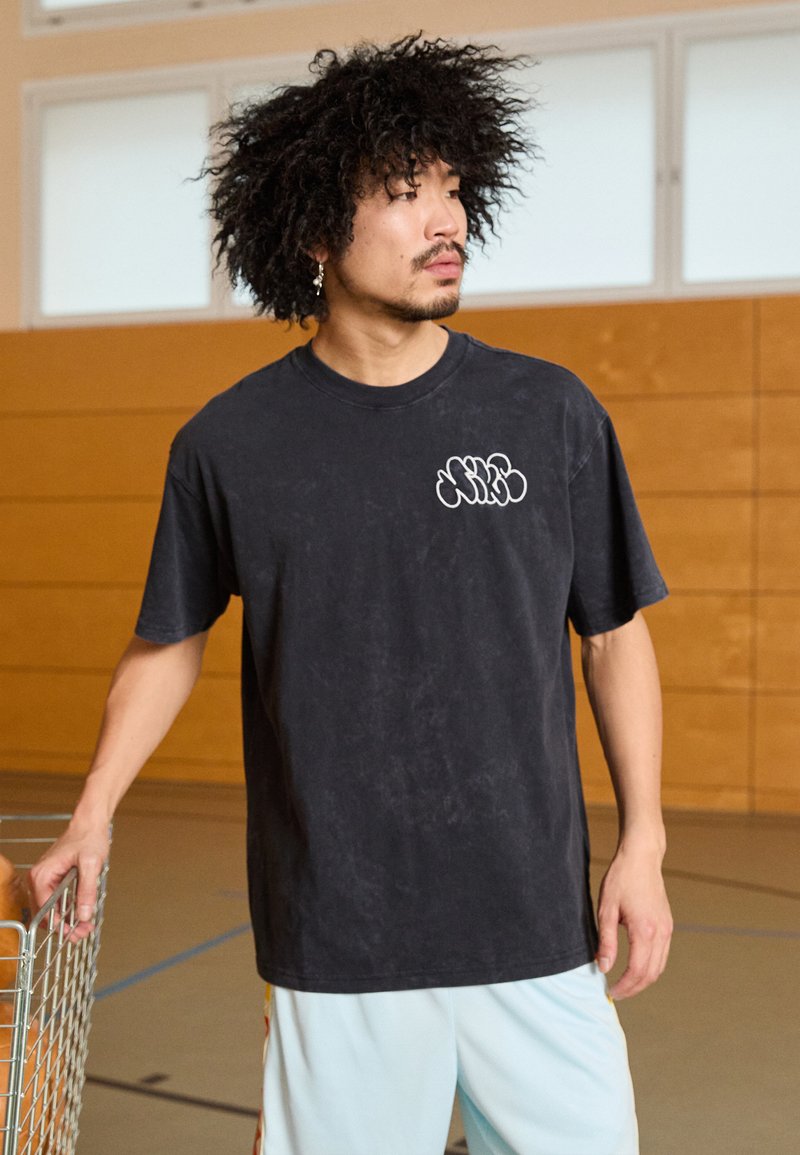 Camiseta de algodón negra con una textura suave, cuello redondo y mangas cortas. Presenta un logotipo gráfico blanco en el lado izquierdo del pecho.