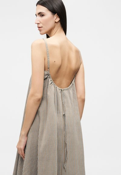 Γυναίκα με μακριά σκούρα μαλλιά που φοράει ένα φαρδύ, αμάνικο μπεζ φόρεμα με λευκές κάθετες ρίγες και χαμηλή, ραμμένη πλάτη.