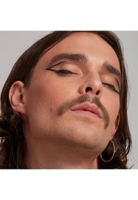 Nahaufnahme eines Gesichts mit einem ausgeprägten Schnurrbart, langen Haaren und einem markanten schwarzen Eyeliner-Design; trägt einen runden Ohrring. Glatte Hautstruktur.