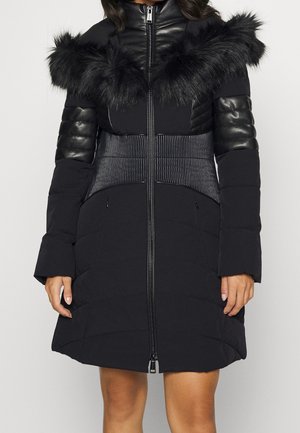 Czarna, pikowana kurtka puchowa do kolan z kołnierzem wykończonym futrem, pikowanymi skórzanymi rękawami i przednim zamkiem błyskawicznym, noszona przez osobę stojącą na jednolitym tle.