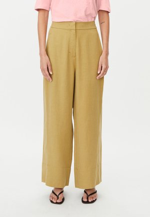 Mujer con pantalones anchos de talle alto en color mostaza, una camisa rosa claro y sandalias negras con los dedos abiertos, de pie sobre un fondo blanco liso.
