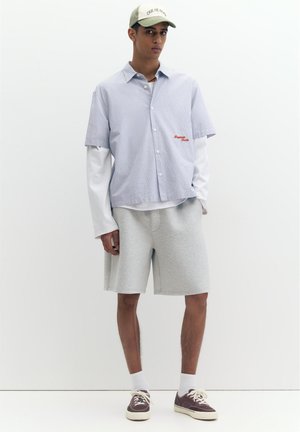 Junger Mann trägt ein hellblaues Kurzarmhemd über langen weißen Ärmeln, graue Shorts, lila Turnschuhe, weiße Socken und eine beige Kappe.