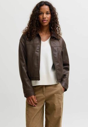 Femme aux cheveux bouclés portant une veste en cuir marron foncé sur un pull blanc et un pantalon beige, debout devant un fond clair uni.