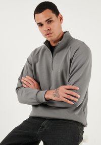 Šedý pulovr se čtvrtinovým zipem vyrobený z hladkého materiálu s raglánovými rukávy, se malým logem na hrudi a uvolněným střihem.