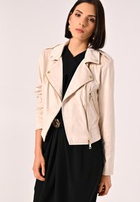 Giacca da moto in pelle beige chiaro con zip frontale, tasche laterali e bottoni a pressione. Presenta un design aderente e maniche lunghe.