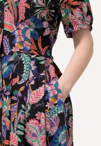 Blumenkleid mit kurzen Ärmeln, das lebendige Farben auf schwarzem Hintergrund, Falten und Seitentaschen bietet. Hergestellt aus leichtem Stoff.