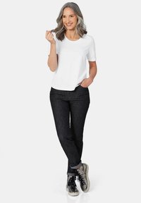 Witte korte mouwen t-shirt gecombineerd met donkere spijkerbroeken, opgerolde mouwen en zilverkleurige sneakers. Model heeft golvend grijs haar.