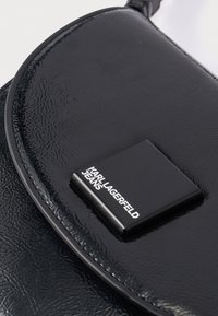 Černá kabelka z umělé kůže s texturovaným povrchem a lesklým povrchem. Má čtvercovou černou plaketu s bílým logem.