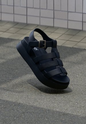 Čierna platformová sandála s prackou na remienku a viacerými vodorovnými remienkami, lietajúca nad textúrovaným chodníkom pri obloženej stene.