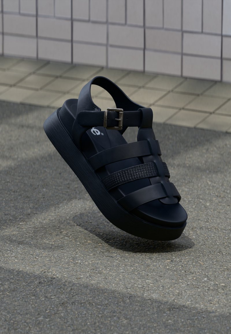 Μαύρο πλατφόρμα σανδάλι με ρυθμιζόμενο λουράκι και πολλαπλές οριζόντιες λουρίδες, αιωρούμενο πάνω από υφή πεζοδρομίου κοντά σε πλακιδωτό τοίχο.