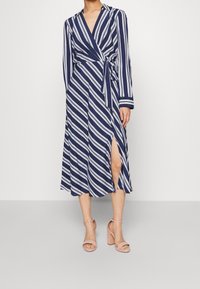 Tmavě modré zavinovací šaty s bílými diagonálními pruhy, dlouhými rukávy, zavazováním v pase a bočním rozparkem. Nošeny s růžovými sandály na blokovém podpatku.