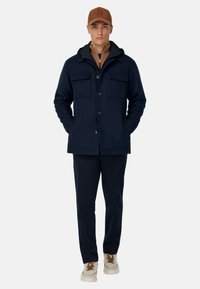 Middengrijze wollen blazer met twee voorkruiszakken en knoopsluiting, gecombineerd met bijpassende broeken en crèmekleurige schoenen met bruine veters.