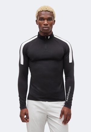 Rövid szőke hajú férfi fekete, hosszú ujjú sportfelsőt visel fehér csíkokkal és logóval az ujjon, fehér nadrággal párosítva, előre néz.