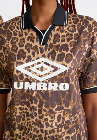 Asmuo vilki rudą ir juodą leopardų rašto sportinę marškinėlį su juodu apykakle ir dideliu baltu "Umbro" logotipu ant krūtinės.