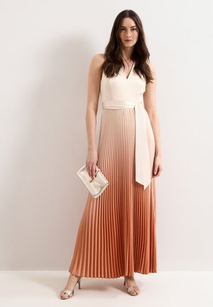 Femme portant une robe maxi plissée beige et pêche sans manches, tenant une petite pochette métallique et portant des sandales beiges à talons hauts.