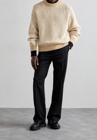 Mann in beigem Strickpullover, schwarzen Hosen und schwarzen Schuhen, der vor einem einfarbigen hellgrauen Hintergrund steht.
