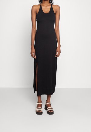 Woman wearing a sleeveless black maxi dress with side slit, paired with white chunky strap sandals, standing against a plain light background.