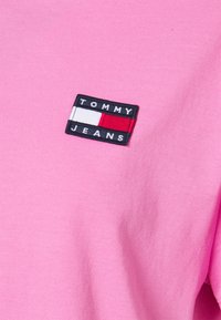 Vaaleanpunainen puuvillapaidat, jossa on suorakulmainen musta läppä valkoisilla ja punaisilla yksityiskohdilla. Läpässä on teksti "TOMMY JEANS" valkoisilla kirjaimilla.