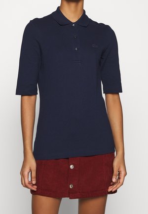 Persona con una camisa polo azul marino de manga corta y una falda de pana roja con botones delanteros sobre un fondo liso.