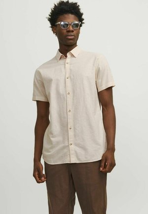 Beige Leinenhemd mit kurzen Ärmeln, Knopfleiste, taillierter Passform und subtiler Textur, ergänzt durch hellbraune Hose.