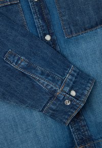 Camicia di denim con un gradiente di colore blu, caratterizzata da bottoni a pressione argentati, cuciture evidenti e un tessuto testurizzato con tonalità di blu variabili.