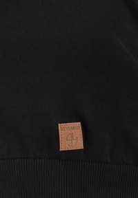 Maglione nero a maglia con orlo a costine e una toppa in pelle marrone texturizzata con testo inciso nell'angolo in basso.