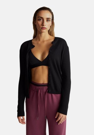 Zwarte cardigan met een ronde hals, voorzien van knoopsluitingen, geribbelde manchetten en een zachte textuur, gecombineerd met een zwarte bh en bordeauxrode broek.