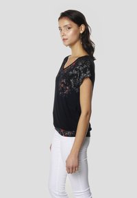 Schwarzes kurzärmeliges Oberteil mit spitzenartigem roten und grauen Muster, V-Ausschnitt-Design und einem kontrastierenden gemusterten Saum, kombiniert mit einer weißen Hose.