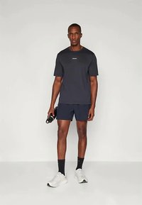 Homem de pé com uma camisola atlética preta e calções pretos, segurando uma garrafa de água preta, usando meias pretas e ténis brancos, em fundo claro.