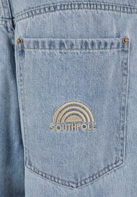 Lyseblå denimlomme med en gylden brodering av en regnbue og ordet "SOUTHPOLE" under. Synlige sømmer og detaljer med metallriveter.