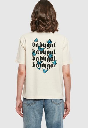Kremfarget t-skjorte med grafisk design som har gjentatt "babygal" tekst og spredte blå sommerfugler på baksiden. Korte ermer.