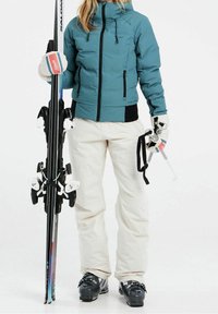 Türkisfarbige Pufferjacke mit schwarzen Akzenten, cremefarbene Skihosen, schwarze Skischuhe, hält Skier mit weißen Bindungen und Stöcken. Strukturierte Handschuhe.