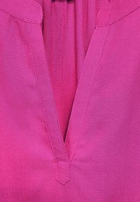 Rosa blus med V-urringning och plisserad design. Slät tyg med synliga sömnadsdetaljer längs med U-ringningen och sidsömmarna.