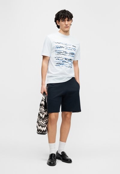 Ανοιχτό μπλε βαμβακερό t-shirt με κυματιστά μπλε μοτίβα, συνδυασμένο με σκούρο μπλε σορτς και μαύρη και λευκή τσάντα με ζιγκ-ζαγκ σχέδιο. Μαύρα loafers και λευκές κάλτσες.