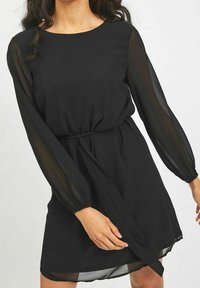 Robe noire avec des manches longues en transparence, encolure ronde, taille cintrée avec un lien, et un ourlet légèrement évasé, mettant en valeur un tissu léger.