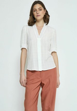 White blouse with short puff sleeves, textured check pattern, and frilled collar detailing, paired with rust-colored wide-leg trousers.