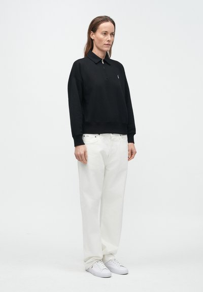 Svart kort genser med glidelåshals, kombinert med løse hvite bukser og hvite joggesko. Enkle, ensfargede plagg uten synlige mønstre eller detaljer.