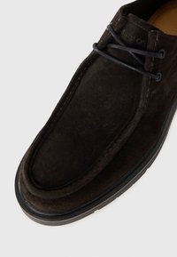 Zapato de ante marrón con punta redondeada, que presenta cordones oscuros, un borde cosido y una suela de goma negra en contraste.