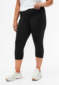 Črne kapri leggins z gladko teksturo in elastičnim pasom, v kombinaciji s svetlo sivo, teksturirano športno obutvijo.