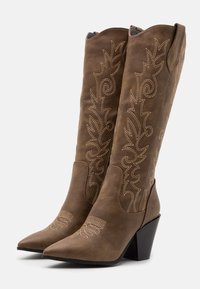 Hnědé semišové kovbojské boty s špičatou špičkou, texturovaným šaftem a bílými vyšívanými květinovými vzory. Mají masivní černý podpatek.