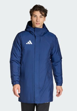 Joven con una chaqueta larga acolchada azul con capucha y logo blanco, de pie frente a un fondo claro y liso.