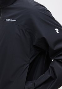 Εγγύς λήψη του μανικιού και του ώμου ενός μαύρου μπουφάν με το λευκό λογότυπο "Peak Performance" στο στήθος και γεωμετρικό λογότυπο στο πάνω μέρος του βραχίονα.