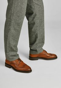 Braune Lederbrogues mit perforierten Details, kombiniert mit grauen strukturierten Hosen, die eine schmal zulaufende Passform und umgeschlagene Bündchen haben.