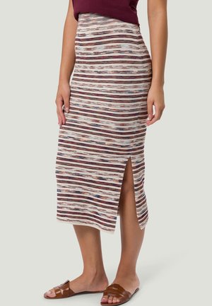 Mujer con una falda midi a rayas con una abertura frontal, un top burdeos y sandalias planas marrones, de pie frente a un fondo liso.