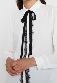 Camicia bianca con bordo in pizzo nero e fiocco al colletto. Maniche lunghe con polsini con bottoni e dettagli delicati lungo la parte anteriore.