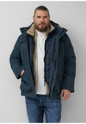 Baardige man die een gelaagde marineblauwe winterjas met fleece voering draagt, over een beige vest met rits en een wit T-shirt, staand tegen een grijze achtergrond.