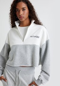 Grijze en witte cropped zip-up sweatshirt met lange mouwen, met "San Francisco" geborduurd in donkerblauw. Zachte textuur en een ontspannen pasvorm.
