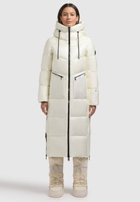 Cappotto lungo e voluminoso di colore bianco con colletto alto e dettagli in cerniera nera. Presenta tasche laterali e un tessuto leggero e lucido, abbinato a stivali beige.