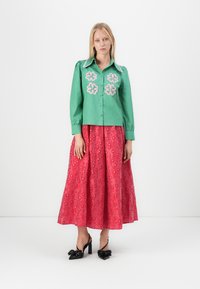 Camisa verde de botones con bordado floral, combinada con una falda rosa hasta la rodilla con patrones florales. Zapatos de tacón negro.